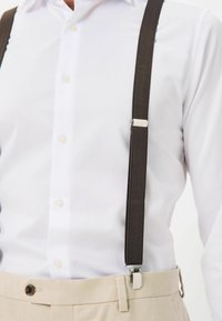 Barna felfüggesztők ezüst csatokkal, fehér inggel és bézs nadrággal párosítva. A felfüggesztők textúrája szőtt.