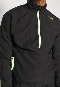 Svart dragkedja med hög krage, neongrön dragkedja och detaljer, med en kenguruficka och en lätt, slät textur.