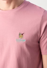 T-shirt en coton rose à manches courtes avec un petit verre à boire brodé et le texte "Lindbergh" en bleu clair en dessous.