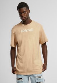 Beige oversized t-shirt with short sleeves, featuring a white printed logo on the chest. Worn with distressed light blue jeans.