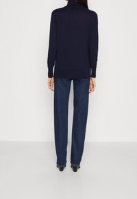 Vrouw die met haar rug naar de camera staat, gekleed in een marineblauwe coltrui, rechtvallende donkerblauwe jeans en zwarte hakken, tegen een effen achtergrond.