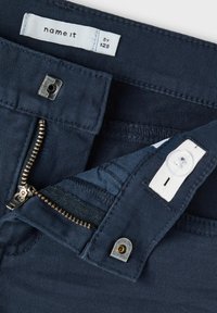 Pantaloni blu navy con chiusura a zip e bottone, realizzati in un tessuto testurizzato e con etichetta in vita. Dettagli di cucitura visibili e accenti in metallo.