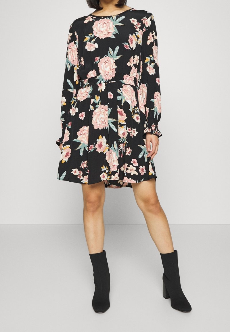 Robe noire à fleurs avec des manches longues, ornée de fleurs roses et bleues. Le tissu texturé se drape en une jupe plissée, accompagnée de bottines noires.