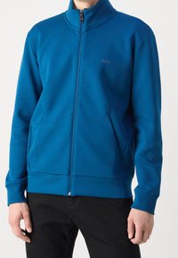 Blå ziptröja i slät tyg, med hög krage, sidofickor och liten logotypdetalj på bröstet. Svarta byxor synliga.