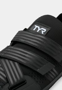 Črne športne čevlje z tremi črtastimi Velcro trakovi, teksturiran material in izstopajoč logo TYR na zgornjem delu.