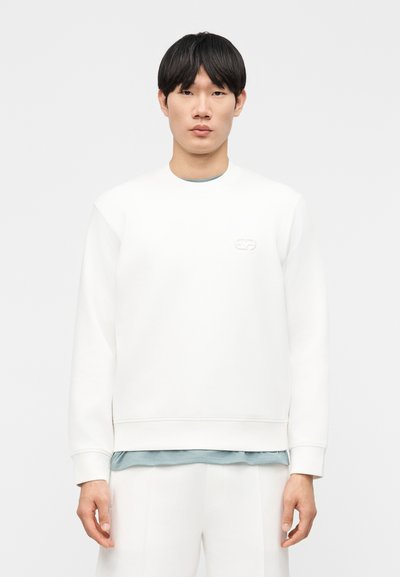 Jeune homme aux cheveux courts et noirs portant un sweat-shirt blanc à manches longues par-dessus une chemise bleu clair, debout devant un fond uni.