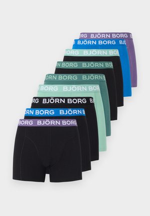 Μυώδης άνδρας κάθεται σε χαλί, στηρίζοντας το κεφάλι του στο χέρι, φοράει μαύρα μπόξερ Björn Borg σε απλό λευκό φόντο.