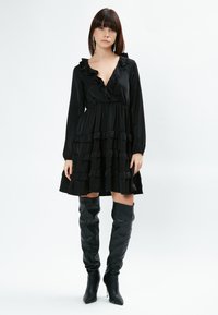 Vestido negro rizado con mangas largas y falda en capas; combinado con botas negras ajustadas hasta el muslo con puntera puntiaguda.