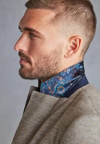 Geknöpfter Blazer aus strukturiertem braunem Stoff, mit einem gemusterten Kragen in Blau und Türkis sowie floralen Details, getragen im Profil.