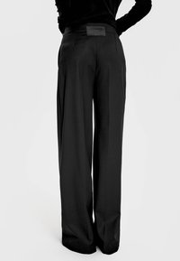 Dopasowane czarne spodnie o szerokim kroju, gładka tekstura i subtelna skórzana łatka z tyłu. Prosty i klasyczny krój.