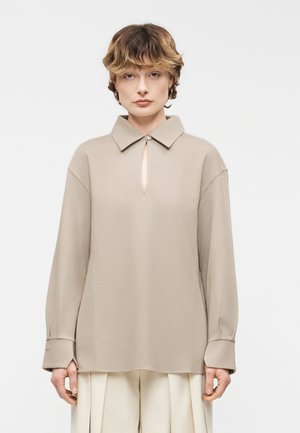 Mladý človek s krátkymi, vlnitými vlasmi oblečený v béžovej košeli s dlhým rukávom, golierom a jedným gombíkom, v kombinácii s svetlými plisovanými nohavicami.