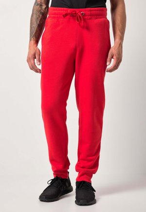 Homme portant un pantalon de survêtement rouge vif avec un cordon de serrage et des chaussures de sport noires, debout devant un fond clair uni.