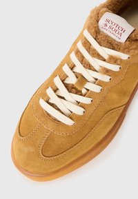 Baskets en daim beige avec lacets blancs, doublure pelucheuse, semelle en gomme et étiquette "Scotch & Soda Amsterdam" sur la languette.