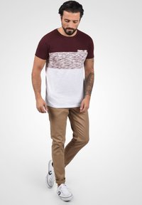 Kortærmet T-shirt med en bordeaux farvet overdel, der har en struktureret hvid vandret stribe og lomme; sat sammen med beige bukser og hvide sneakers.