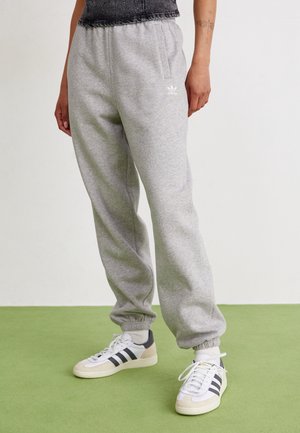 Osoba w jasnoszarych spodniach dresowych Adidas i białych butach Adidas z czarnymi paskami stojąca na zielonej podłodze.