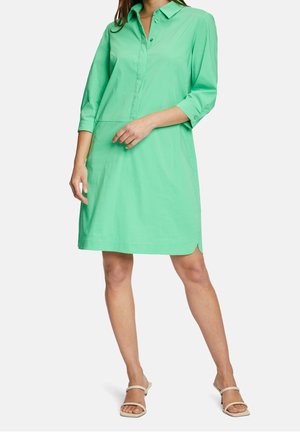 Femme portant une robe chemise mi-longue vert menthe avec des manches trois-quarts et des sandales à talons beige à bout ouvert sur fond blanc.