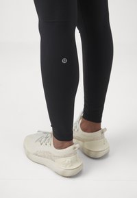 Czarne prążkowane legginsy z małym logo przy kostce, połączone z białymi butami sportowymi o wzorzystym designie i wyraźnym obcasie.