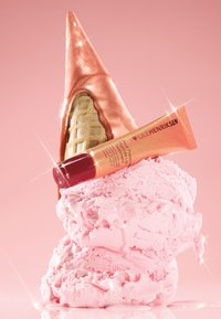 Różowe kulki lodów o błyszczącej teksturze, zwieńczone lśniącym miedzianym rożkiem. Obok znajduje się tubka balsamu do ust w czerwono-złotym kolorze.