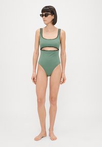 Viengabiegu peldkostīms muted zaļā krāsā ar izgriezumu. Iezīmējas ar kontrastējošu melnu apdari ap apkakli un roku atverēm. Gluda tekstūra.