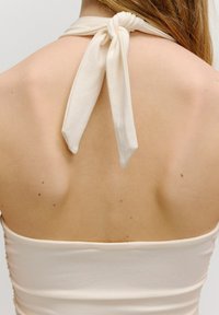 Crème-kleurige haltertop met een gekruiste nekband aan de achterkant, gladde textuur en aansluitende stijl, die de bovenrug en schouders accentueert.
