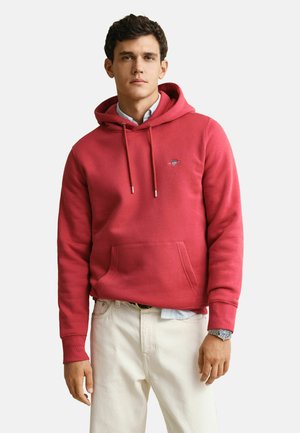 Junger Mann trägt einen roten Kapuzenpullover, hellblaues Hemd, weiße Hose und eine Armbanduhr, steht vor einem schlichten Hintergrund.