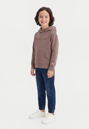 Smilende barn stående iført en brun Champion hættetrøje, mørkeblå jeans og hvide sneakers mod en ensfarvet lys baggrund.
