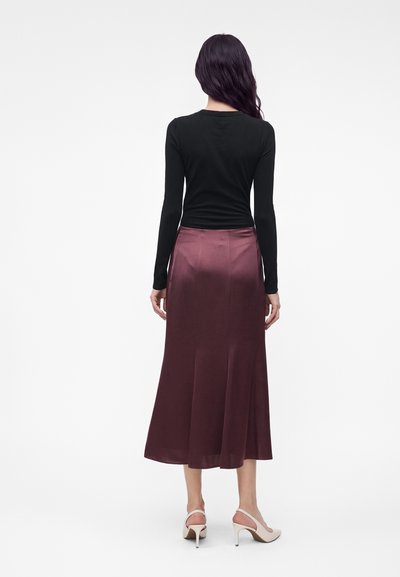 Femme aux longs cheveux foncés portant un haut noir à manches longues, une jupe midi violette foncée brillante et des chaussures blanches à talons hauts, debout de dos.