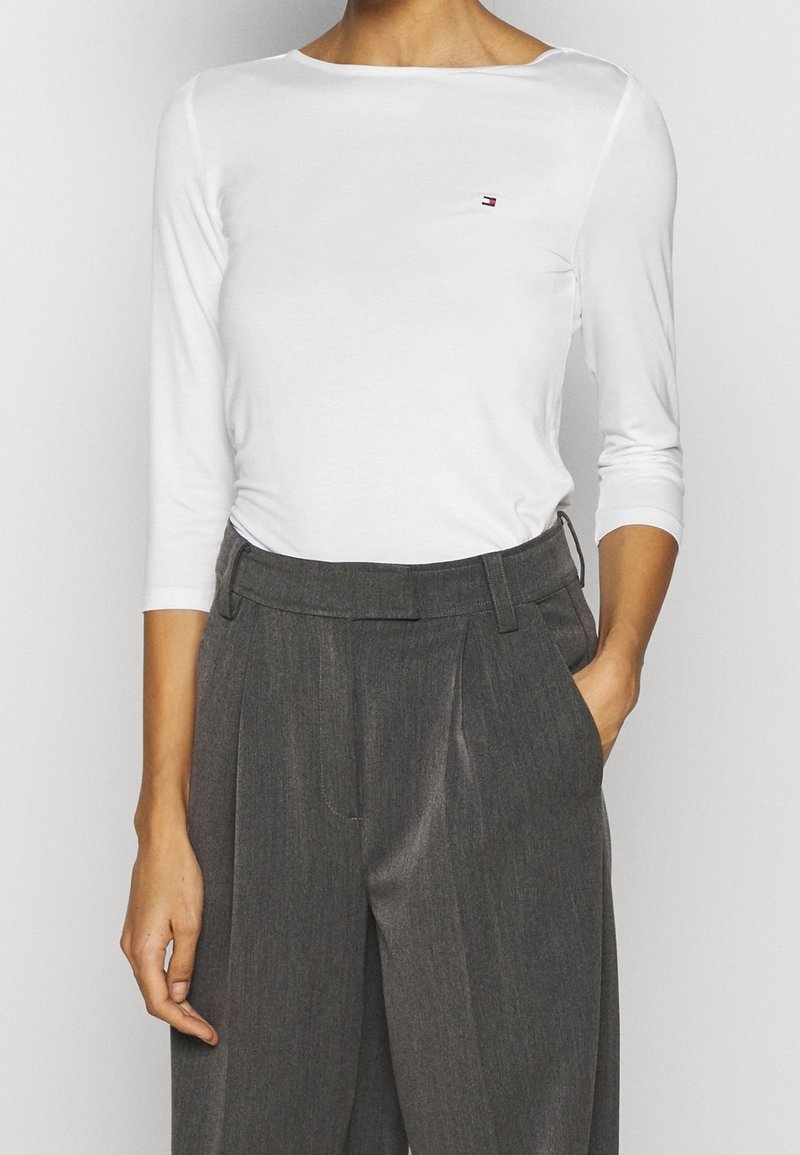 Blusa blanca de manga larga con escote barco y pequeño logo en el pecho, combinada con pantalones grises de tiro alto con pliegues.