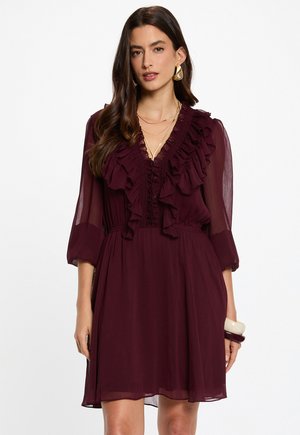 Femme portant une robe bordeaux foncé avec un col en V à volants et des manches trois-quarts en voile, accessoirisée avec des boucles d'oreilles dorées et des colliers superposés.