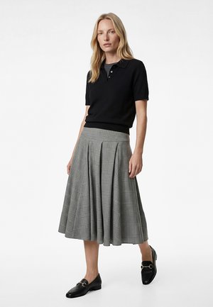 Blonde Frau trägt ein schwarzes Polo-Shirt mit kurzen Ärmeln, einen grauen karierten plissierten Midi-Rock und schwarze Leder-Loafer, steht vor weißem Hintergrund.