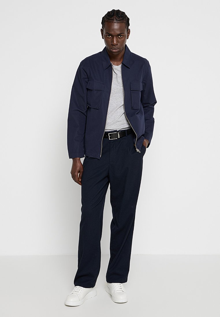 Hombre con trenzas que lleva una chaqueta azul marino, camiseta gris, cinturón negro, pantalones azul marino y zapatillas blancas, de pie contra un fondo blanco.