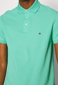 Polo shirt en coton vert avec col, doté d'une patte de boutonnage à trois boutons et d'un petit logo avec des accents rouge, blanc et bleu sur la poitrine.