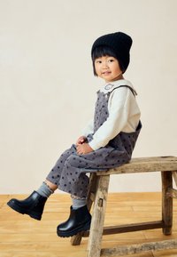 Jeune enfant en salopette à pois, pull blanc, bonnet noir et bottes, assis sur un banc en bois rustique devant un fond uni.
