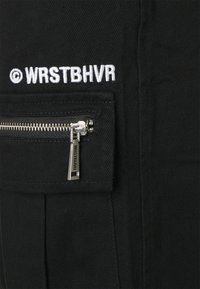Tessuto nero con finitura testurizzata presenta un logo ricamato bianco "© WRSTBVR" sopra una tasca con zip dotata di hardware metallico.