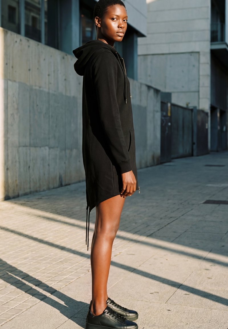 Junge Person trägt ein schwarzes Hoodie-Kleid und schwarze Sneakers und steht auf einem sonnigen städtischen Gehweg neben einer Betonwand.
