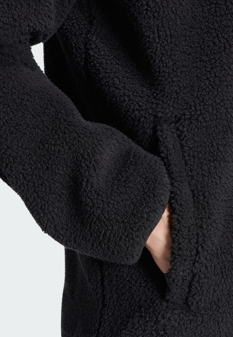 Gros plan d’une main dans la poche d’une veste polaire noire texturée à manches longues.