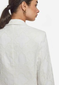 Vit jacka med texturerad tyg och en subtil geometrisk mönstring. Anmärkningsvärda detaljer inkluderar en klassisk krage och skräddarsydd passform. Guld hängande örhänge.