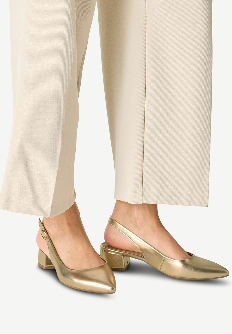 Scarpe slingback metalliche dorate con punta affilata, tacco a blocco e texture liscia, abbinate a pantaloni beige a gamba larga.