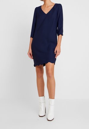 Vestido azul marino con escote en V, mangas de tres cuartos y dobladillo ondulado. Hecho de tela suave, combinado con botas blancas hasta el tobillo.