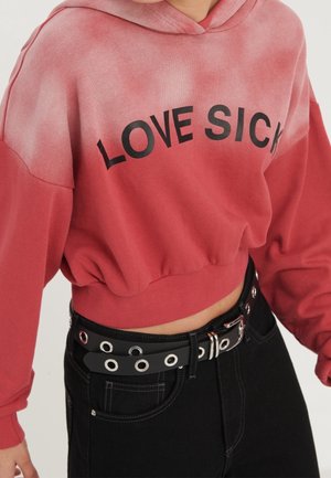 Haut à capuche rouge dégradé, coupé, avec "LOVE SICK" en texte noir. Porté avec une ceinture noire ornée d'œillets en argent et d'une boucle. Jeans noirs visibles.
