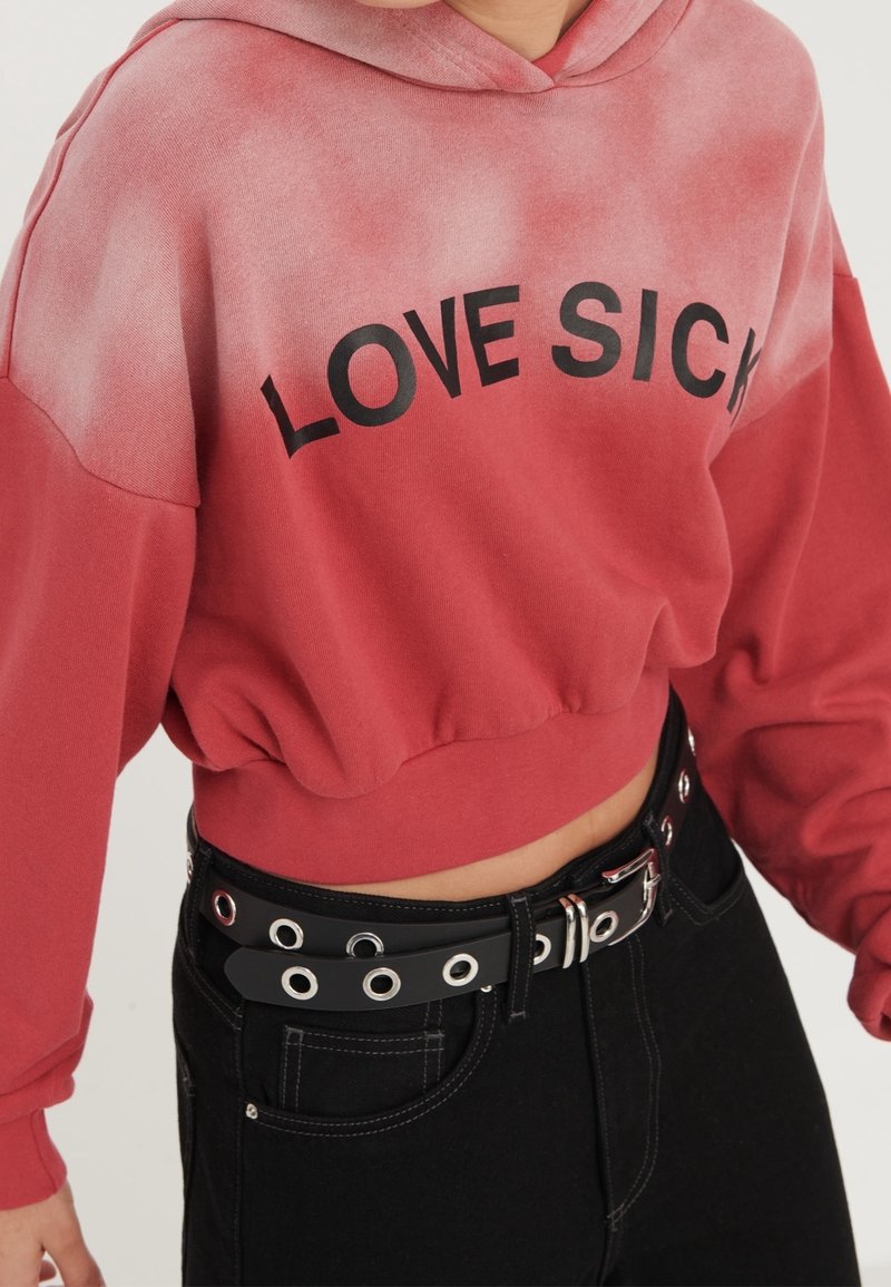 Felpa con cappuccio tagliata in gradiente rosso con la scritta "LOVE SICK" in nero. Indossata con una cintura nera con occhielli in argento e fibbia. Visibili jeans neri.