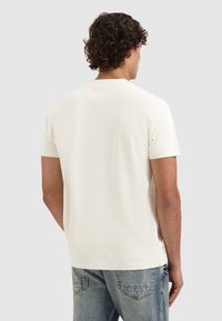 Mann mit lockigem Haar, der ein schlichtes, cremefarbenes kurzärmeliges T-Shirt und blaue Jeans trägt, von hinten auf einem neutralen Hintergrund gesehen.
