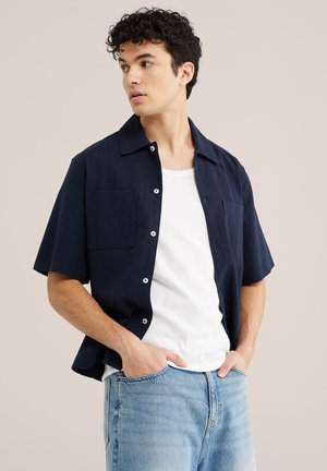 Young man with curly hair wearing open navy short-sleeve shirt over white t-shirt and light blue jeans, looking to the side with hands in pockets.