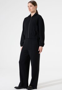 Zwarte ritsjack met elastische mouwen, gecombineerd met hoogtaille zwarte broek met een getailleerde pasvorm en een lichte textuur. Zwarte platte schoenen.
