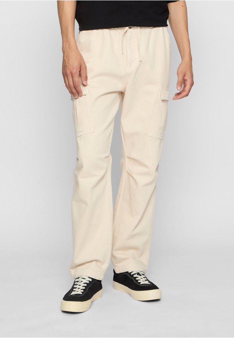 Jasnobeżowe spodnie cargo z elastycznym pasem, dwoma bocznymi kieszeniami i luźnym krojem. Do tego czarne trampki z białymi sznurowadłami.