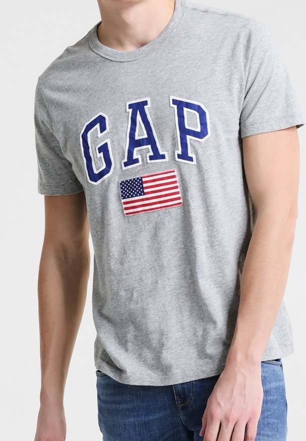 T-shirt gris avec "GAP" en lettres bleues et un petit patch du drapeau américain, porté par une personne avec un arrière-plan neutre.