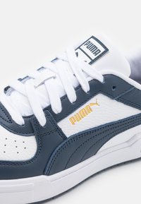 Witte en marineblauwe sneaker met een bovenwerk van getextureerd leer, gouden Puma-logo en witte veters. Voorzien van perforatiedetails en een gevoerde kraag.