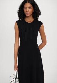 Vestido preto em malha canelada com decote redondo e mangas curtas, apresentando um corpo justo e uma saia larga. A modelo segura uma bolsa branca.