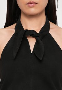 Fekete pántos felsőt viselő nő, akinek a nyakánál megkötött csomó van, jól látható a kulcscsontja, valamint az ajkaiból és az állából egy részt is mutat.