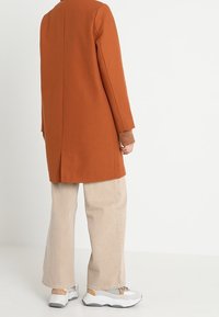 Abrigo largo de color óxido con corte recto, combinado con pantalones de pierna ancha beige y zapatillas grises, que presentan un puño de punto marrón texturizado.